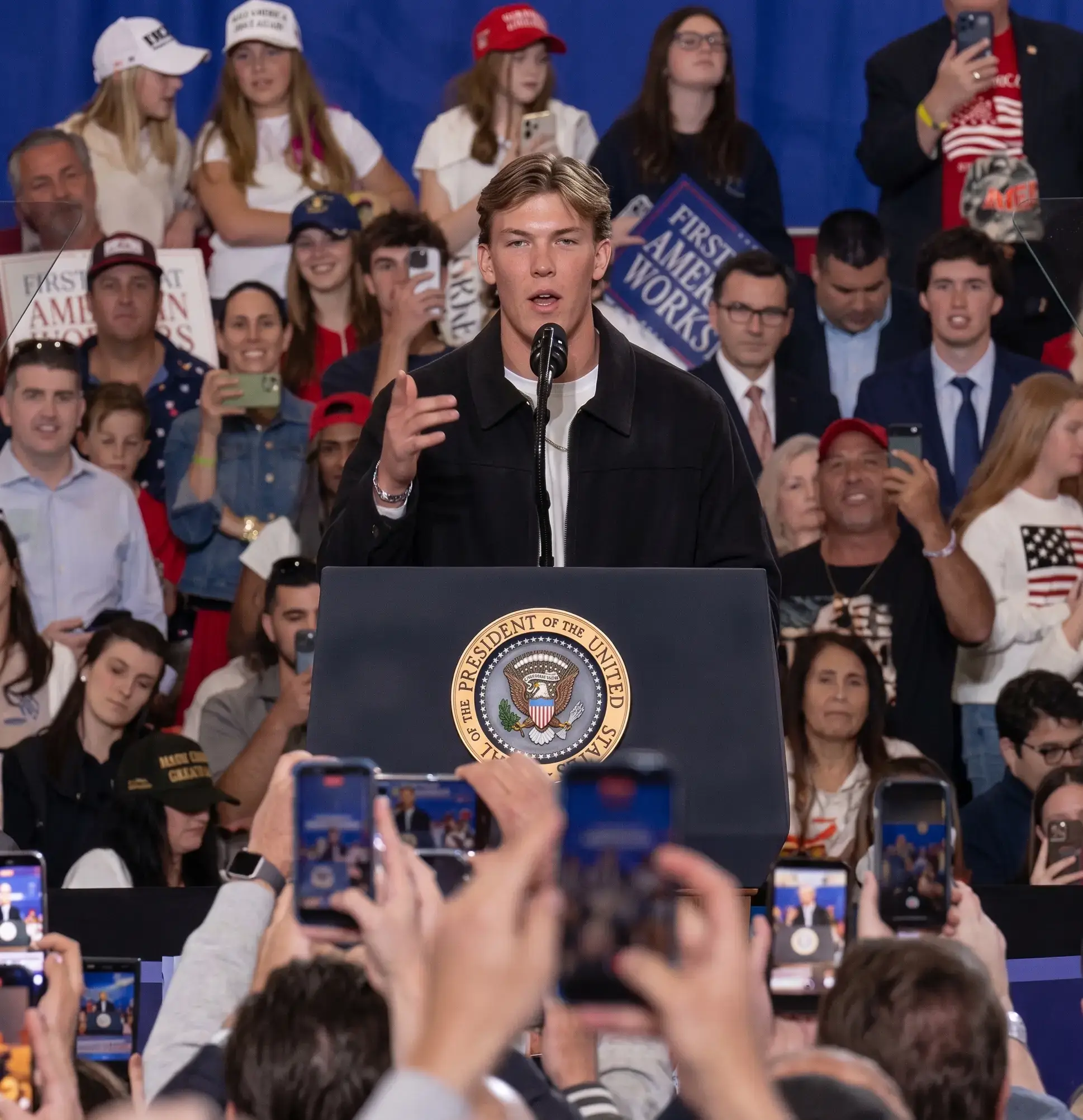Jaxson Dart speaks from a podium bearing the seal of the President of the United States while an audience takes photos with their phones.