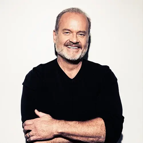 Kelsey Grammer smiling brightly with his arms crossed, wearing a black long-sleeved shirt against a crisp white background.