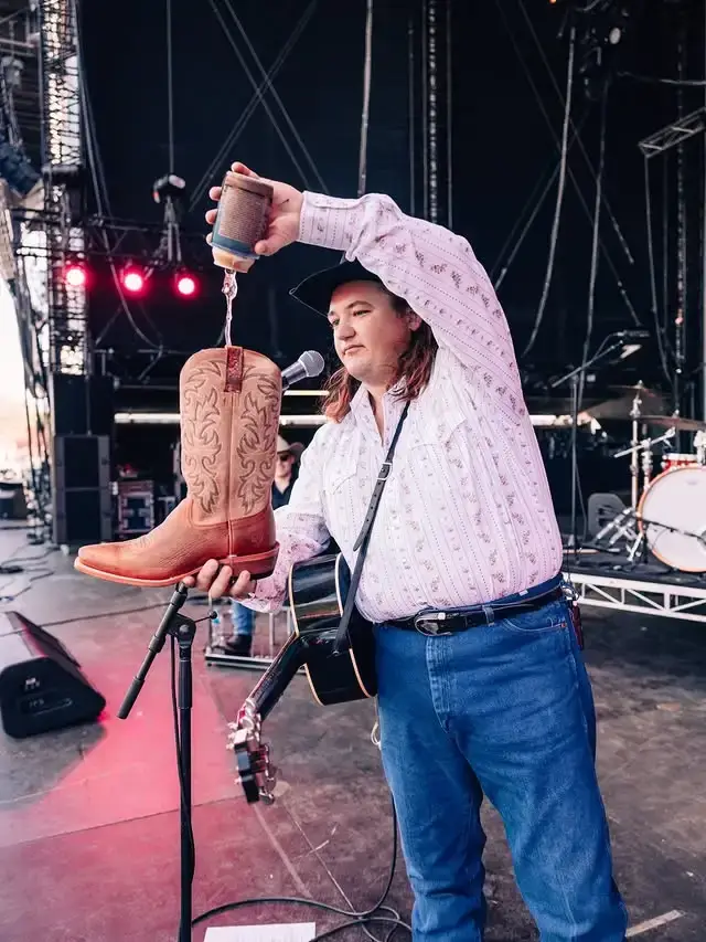Country singer Jake Worthington pours a drink from a can directly into a leather cowboy boot on a concert stage.