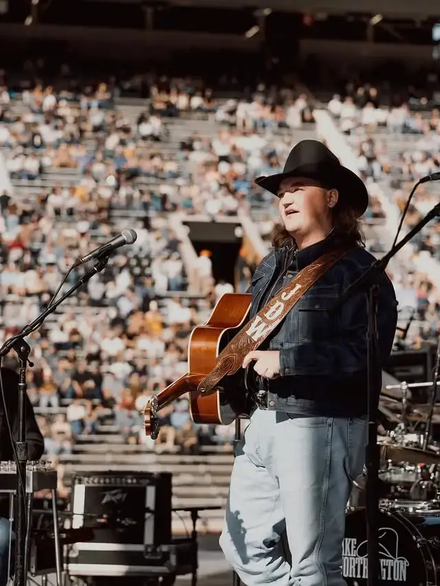 Country artist Jake Worthington plays acoustic guitar on a stadium stage in a cowboy hat and denim jacket in front of a large crowd.