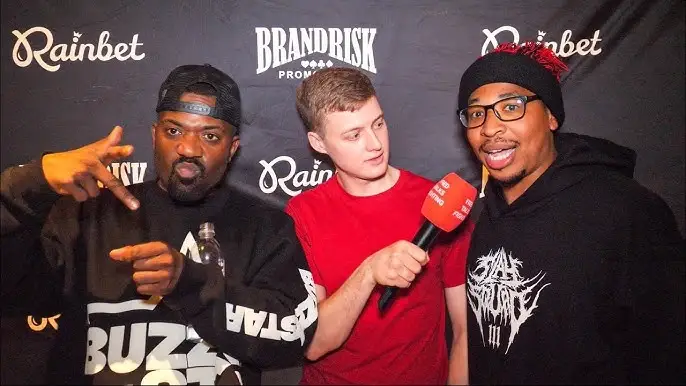 Ray J and internet personality Supa Hot Fire stand on either side of an interviewer holding a red microphone in front of a Brand Risk Promotions backdrop.