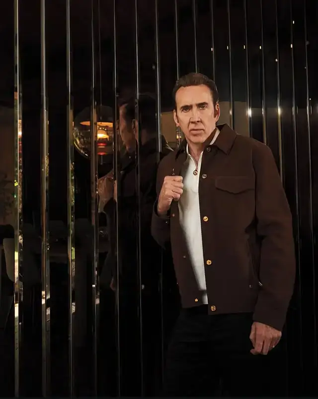 Nicolas Cage stands next to a reflective metallic wall wearing a brown jacket with gold buttons over a white collared shirt.