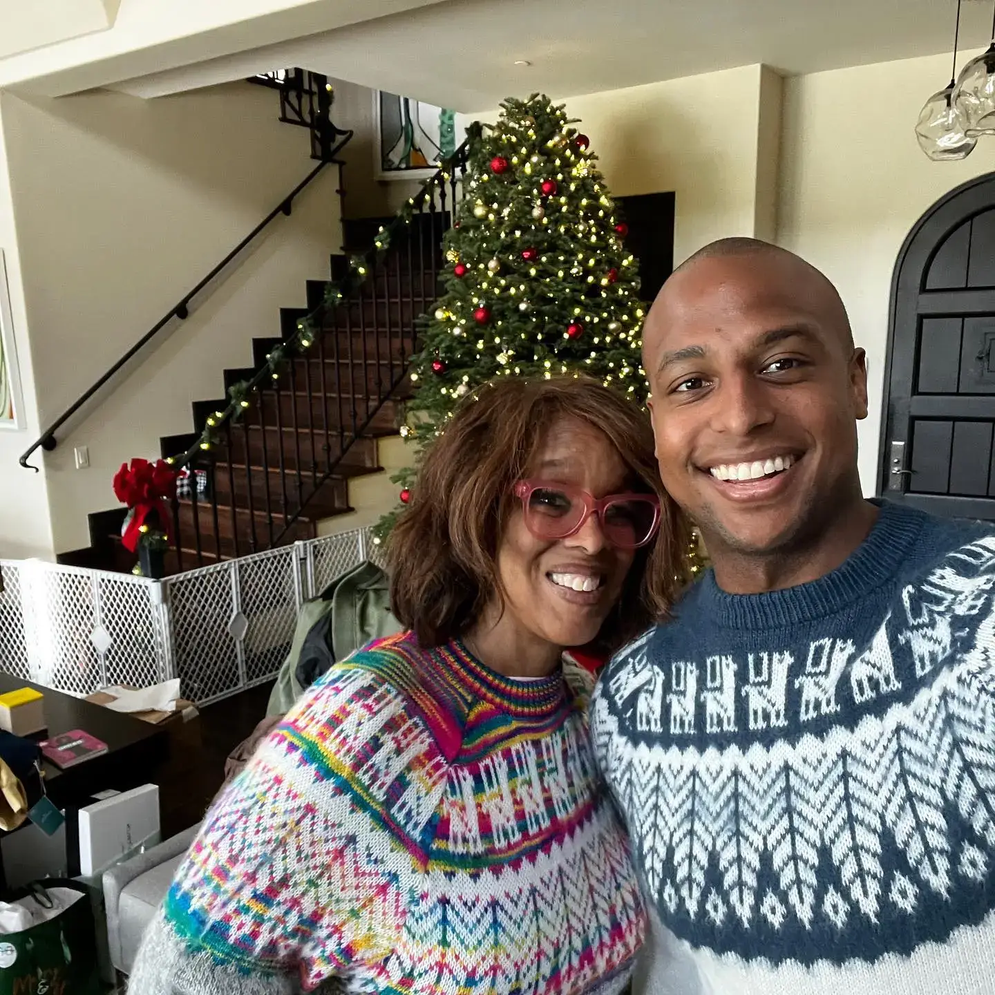 Gayle King in a colorful patterned sweater and pink glasses smiles next to her ex husband, William Bumpus for a holiday selfie in front of a Christmas tree.