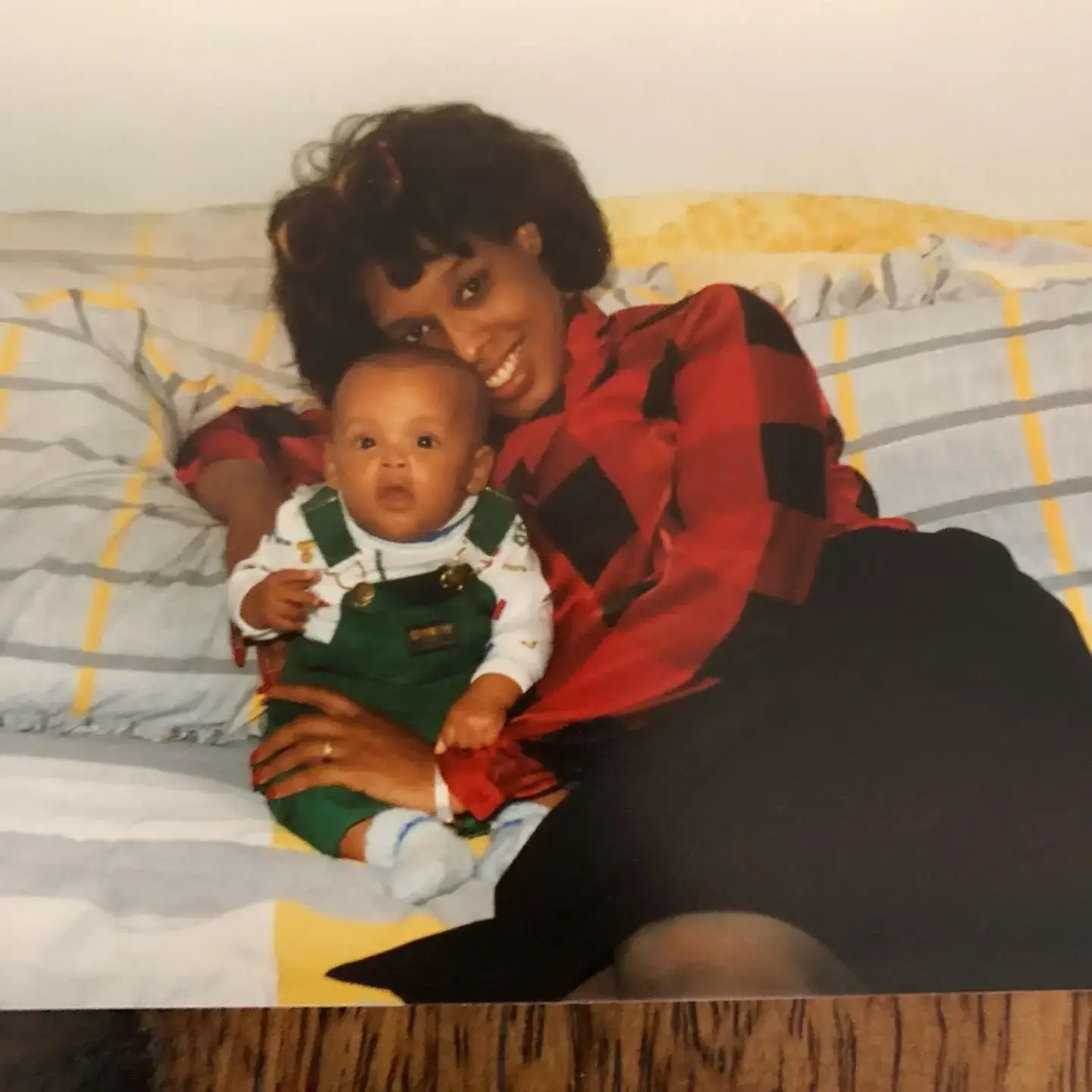 A vintage photograph shows a young Gayle King smiling while holding her infant son, William Bumpus Jr., who is wearing green overalls on a bed.