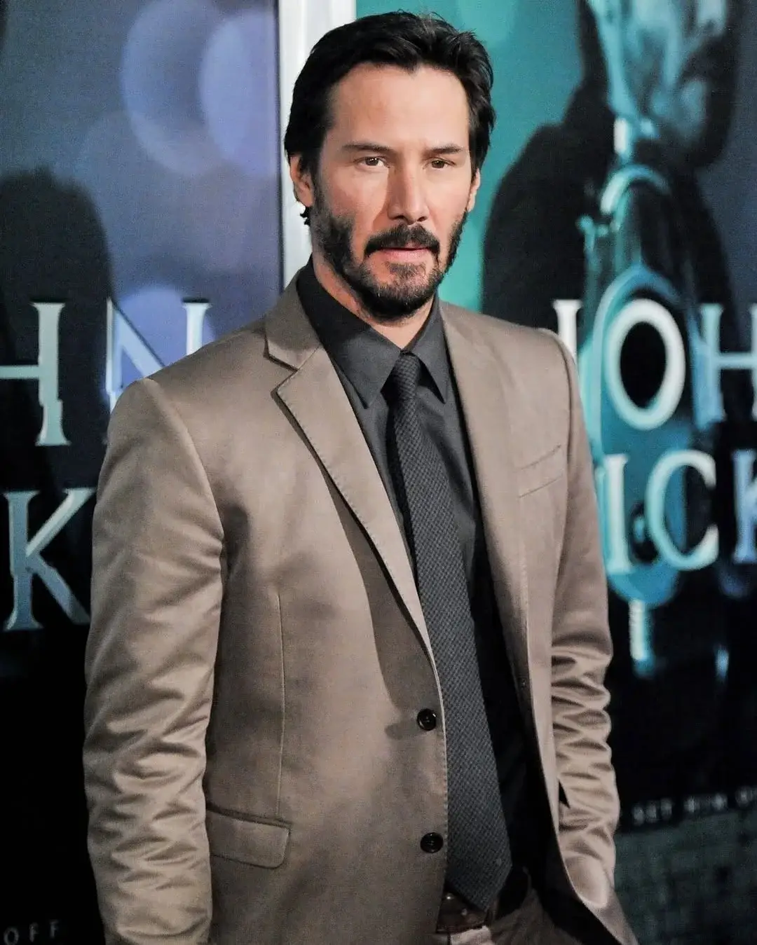 Keanu Reeves poses on a red carpet wearing a tan suit jacket over a dark grey button-down shirt and a matching tie.