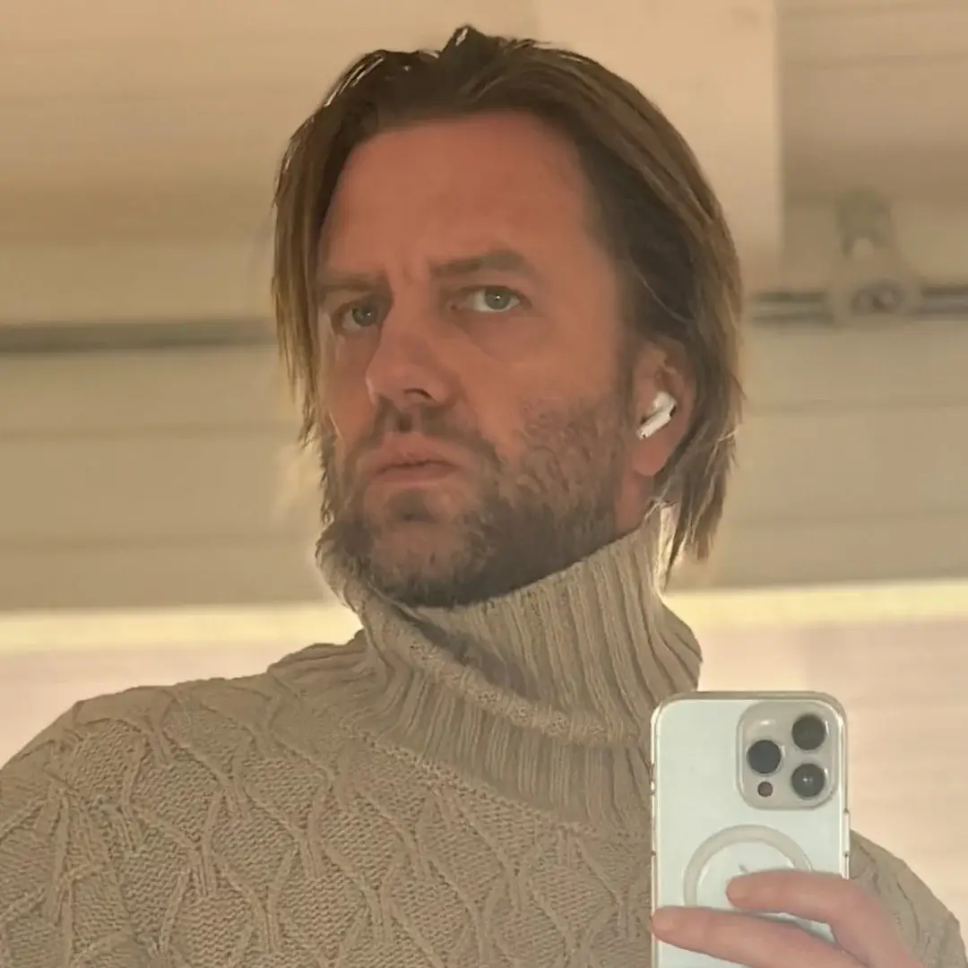 Filmmaker Carl Rinsch takes a mirror selfie while wearing a high-collared tan knit turtleneck sweater and an earbud.
