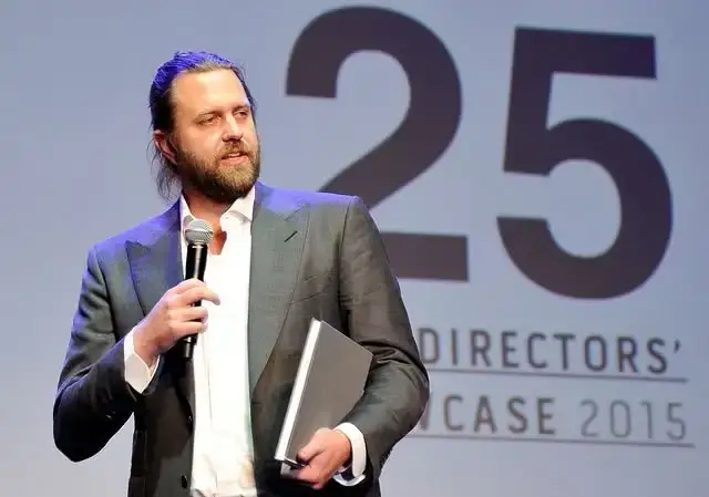 Carl Rinsch stands on a stage holding a notebook and speaking into a microphone in front of a backdrop that reads "25 Directors