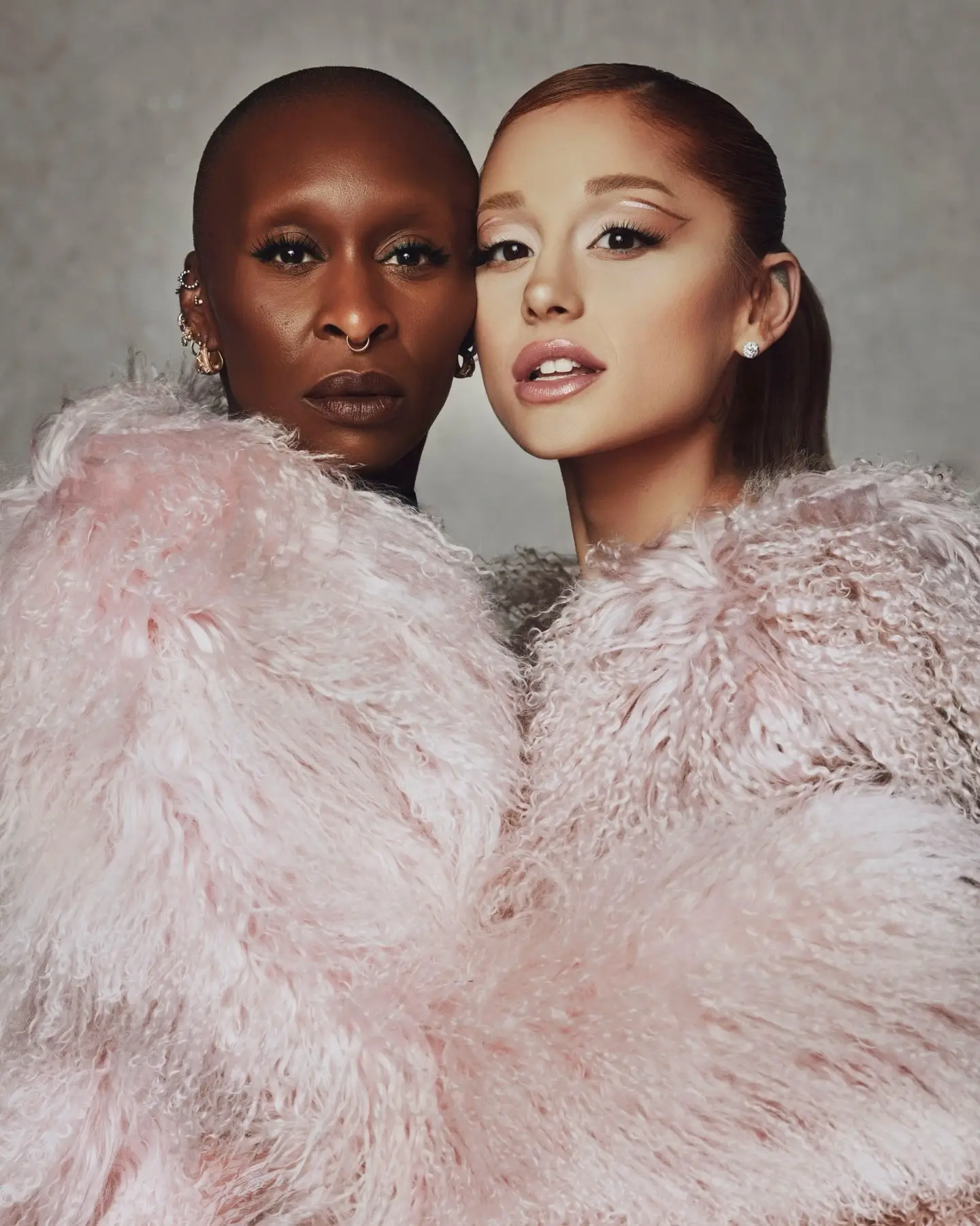 Cynthia Erivo and Ariana Grande pose cheek-to-cheek while wearing matching, voluminous pastel pink textured fur coats against a grey studio backdrop.