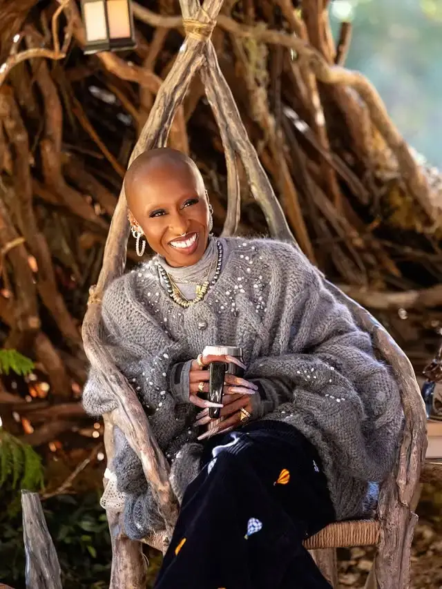 Cynthia Erivo smiles broadly while sitting in a rustic wooden chair wearing a thick, beaded grey knit sweater and holding a travel mug.