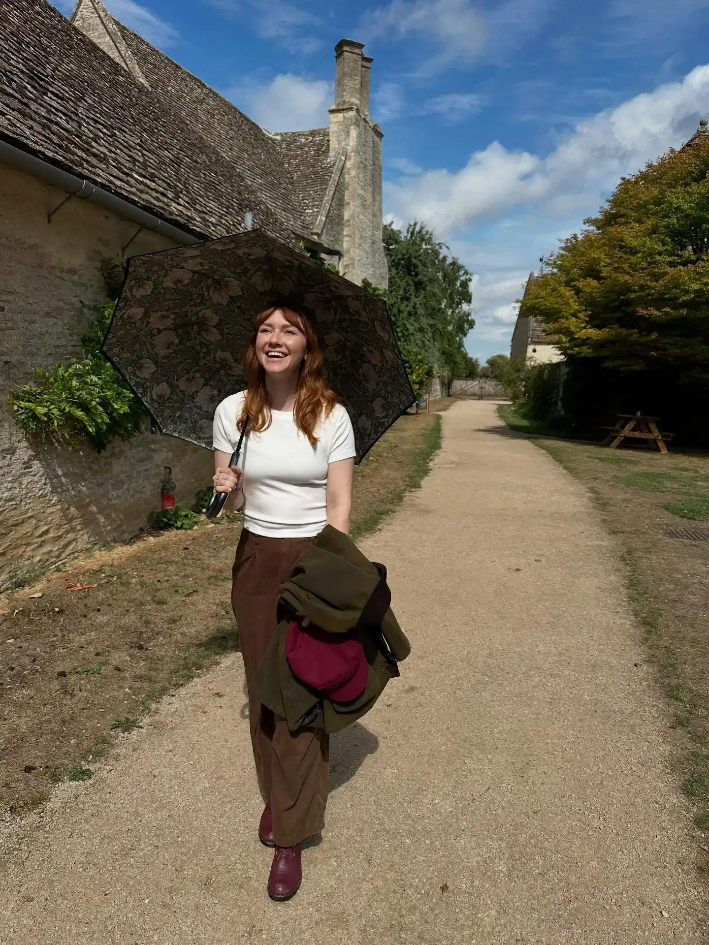 Valorie Curry walks along a dirt pathway while smiling and holding a patterned umbrella next to stone buildings.