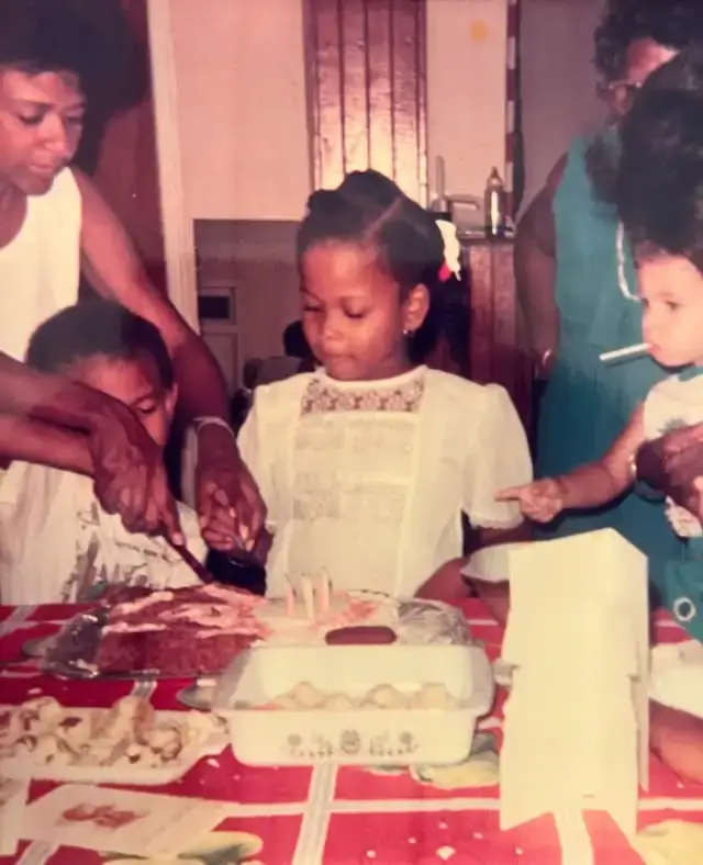 A vintage childhood photo shows a young Nneka Elliott standing behind a birthday cake on a decorated table surrounded by family members.