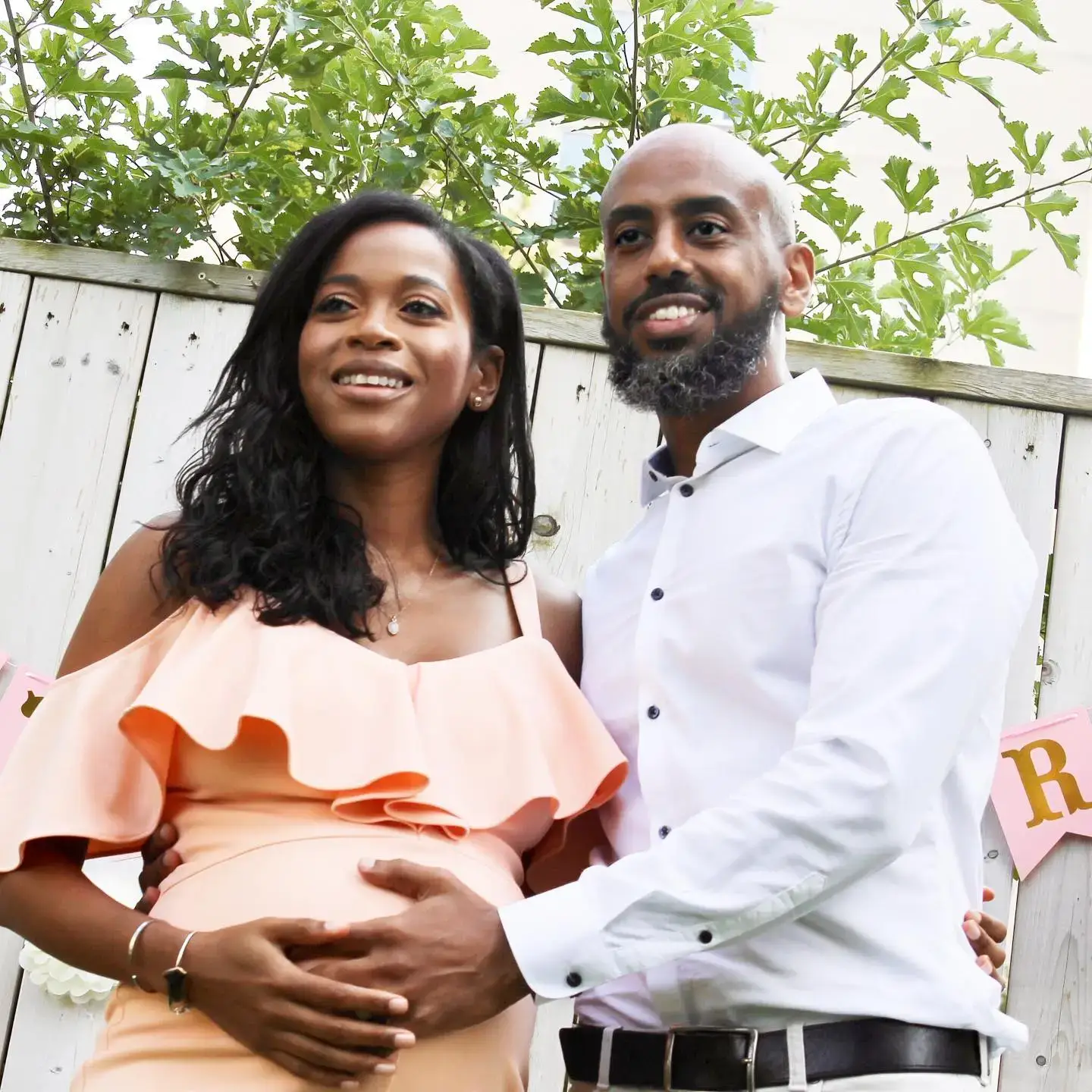 Nneka Elliott in a peach ruffled dress and her husband, Victor Ghebre, cradle her pregnant belly while smiling together in front of a wooden fence.