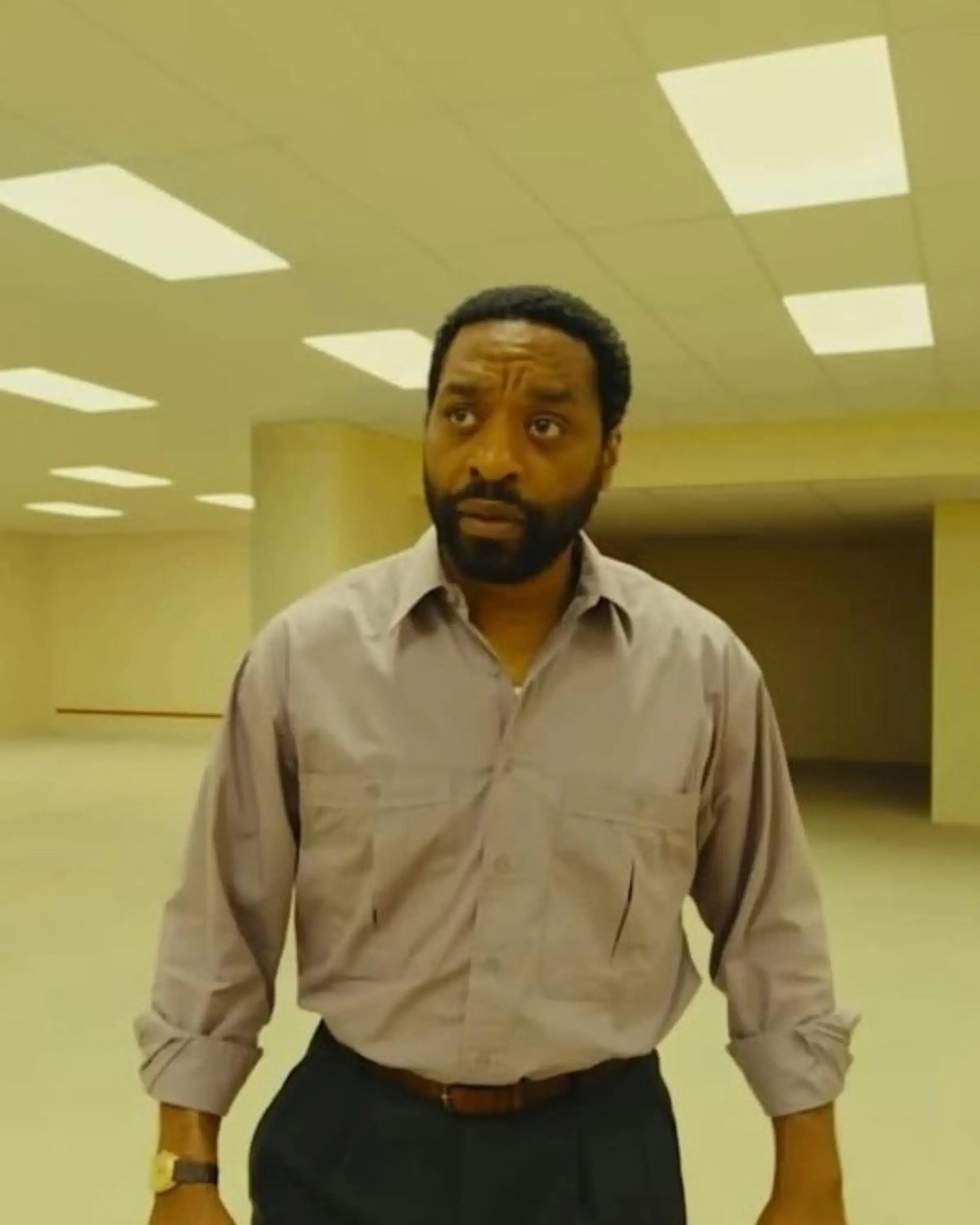 Chiwetel Ejiofor looks around with a concerned and disoriented expression while standing inside a vast, yellow-toned room reminiscent of the Backrooms.