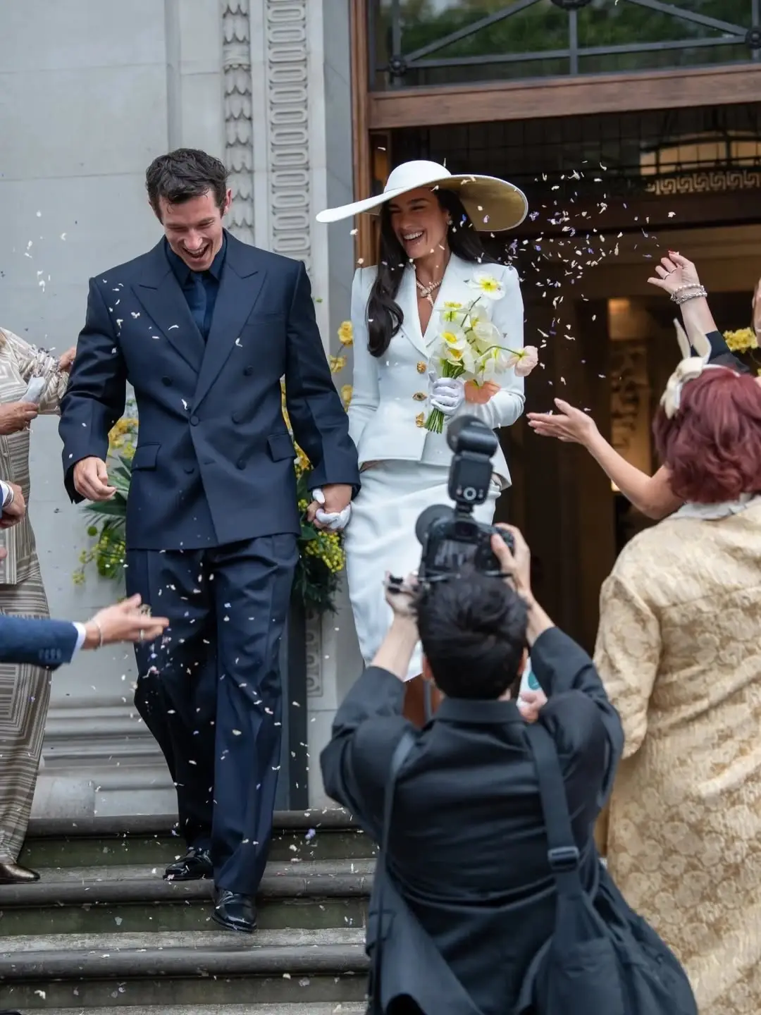 Dua Lipa and Callum Turner hold hands and smile as they exit a building amidst thrown confetti, with Dua in a white suit and wide-brimmed hat and Callum in a dark double-breasted suit.