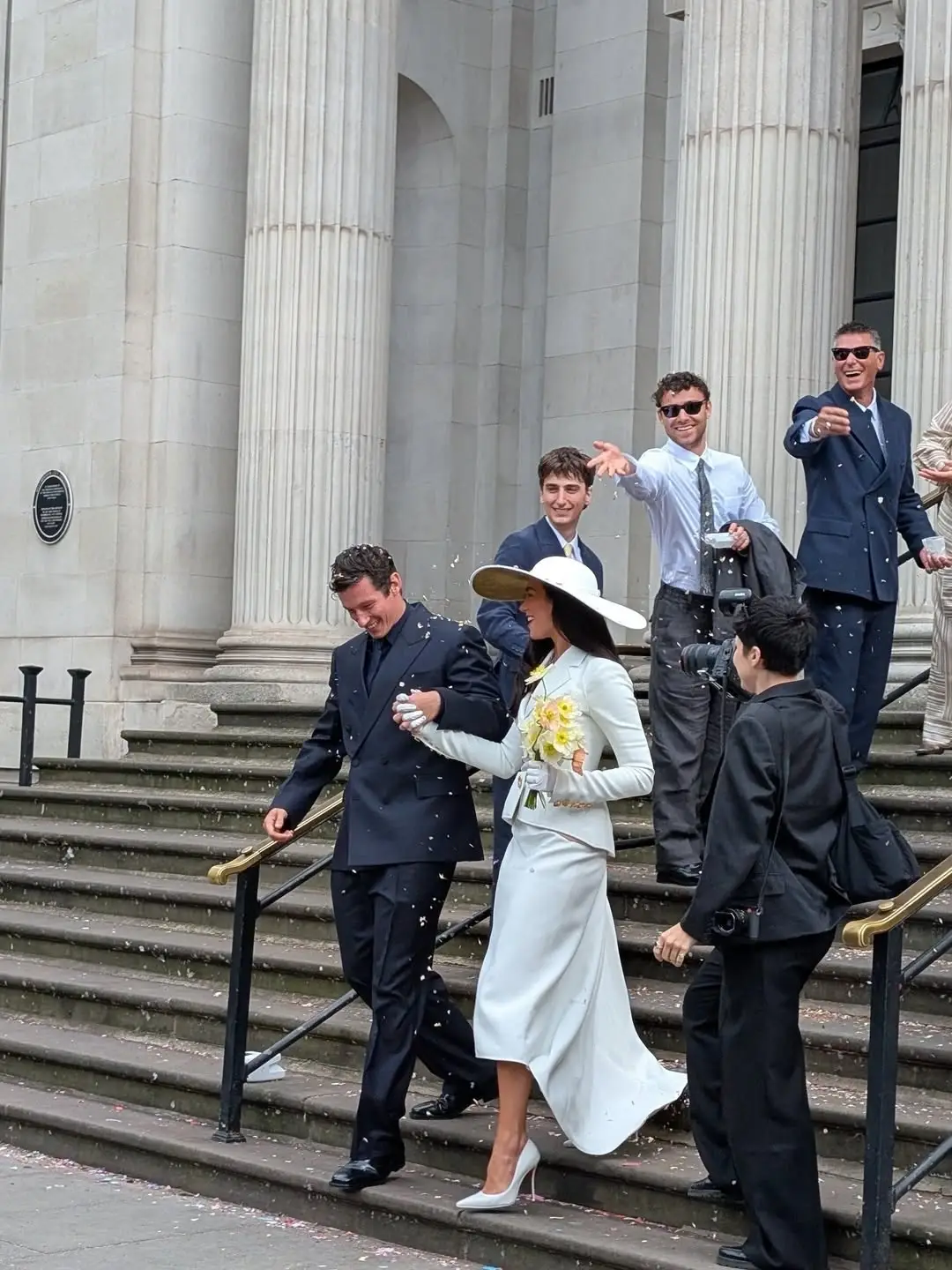 Alt Text: Dua Lipa and Callum Turner hold hands while walking down the steps of a pillared building as guests throw confetti.