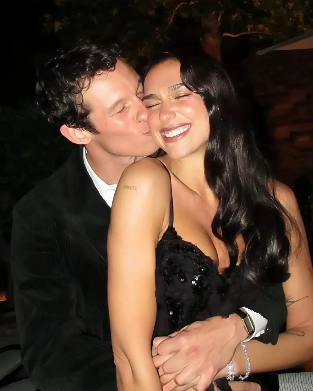 Callum Turner hugs Dua Lipa from behind and kisses her cheek as she smiles brightly in a black sequin dress.