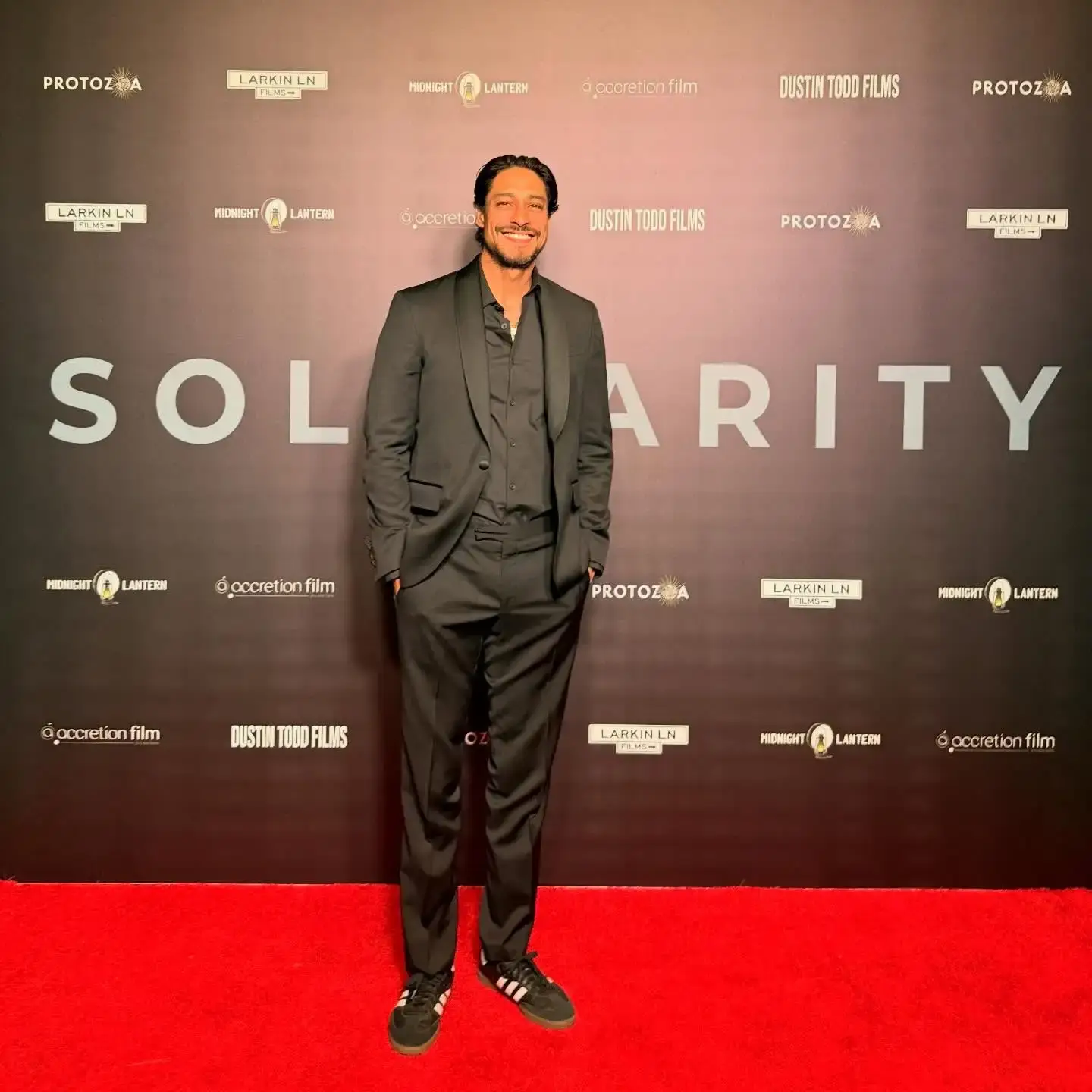 Carlos Miranda stands smiling on a red carpet in an all-black suit and sneakers in front of a backdrop that reads "SOLIDARITY",