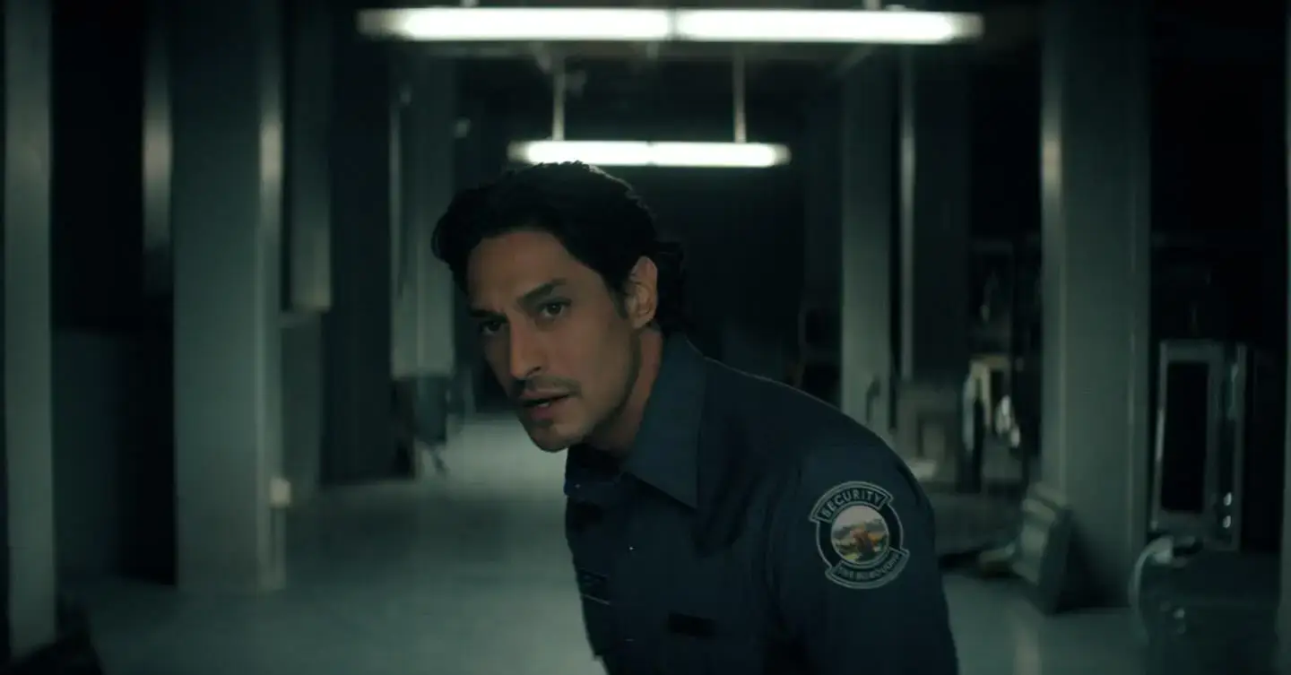 Carlos Miranda wears a dark security guard uniform with a shoulder patch while looking alert in a dimly lit industrial hallway