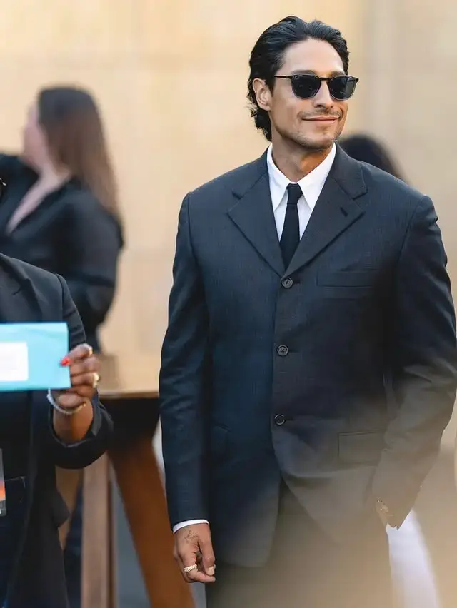 Carlos Miranda smiles slightly while wearing sunglasses, a dark three-button suit jacket, a white collared shirt, and a black tie.