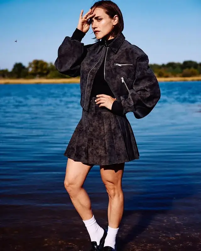Alice Kremelberg poses by the edge of a lake wearing a dark, textured bomber jacket and a matching pleated mini skirt.