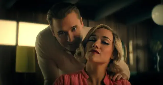 A blonde Alice Kremelberg tilts her head back with a slight smile as a man leans in from behind her with his hand on her shoulder.