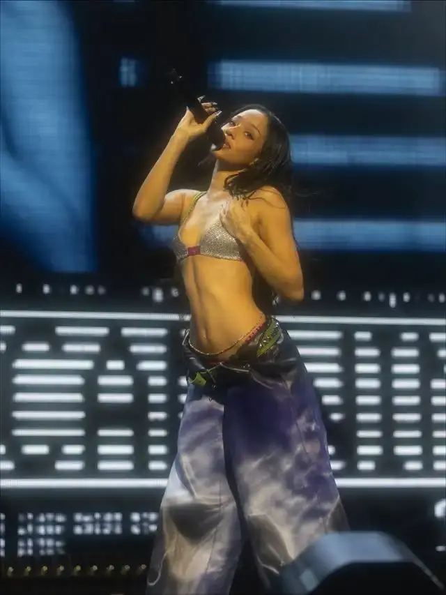A woman performs on a brightly lit stage into a microphone while wearing a metallic silver bralette and oversized patterned trousers.