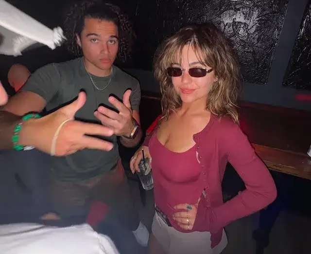 Mika Abdalla wears a reddish-pink top with dark sunglasses while Jalen Thomas Brooks poses next to her making hand gestures in a dimly lit indoor setting.
