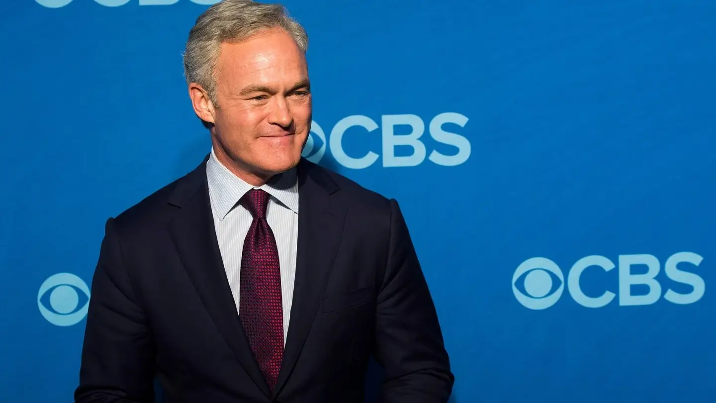 Journalist Scott Pelley smiles while posing in a dark suit and maroon tie against a bright blue CBS backdrop.