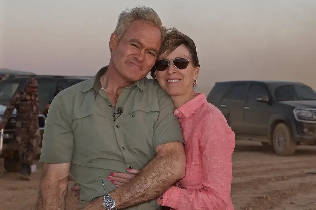Scott Pelley and his wife, Jane, stand close together and smile in an outdoor desert setting with vehicles in the background.