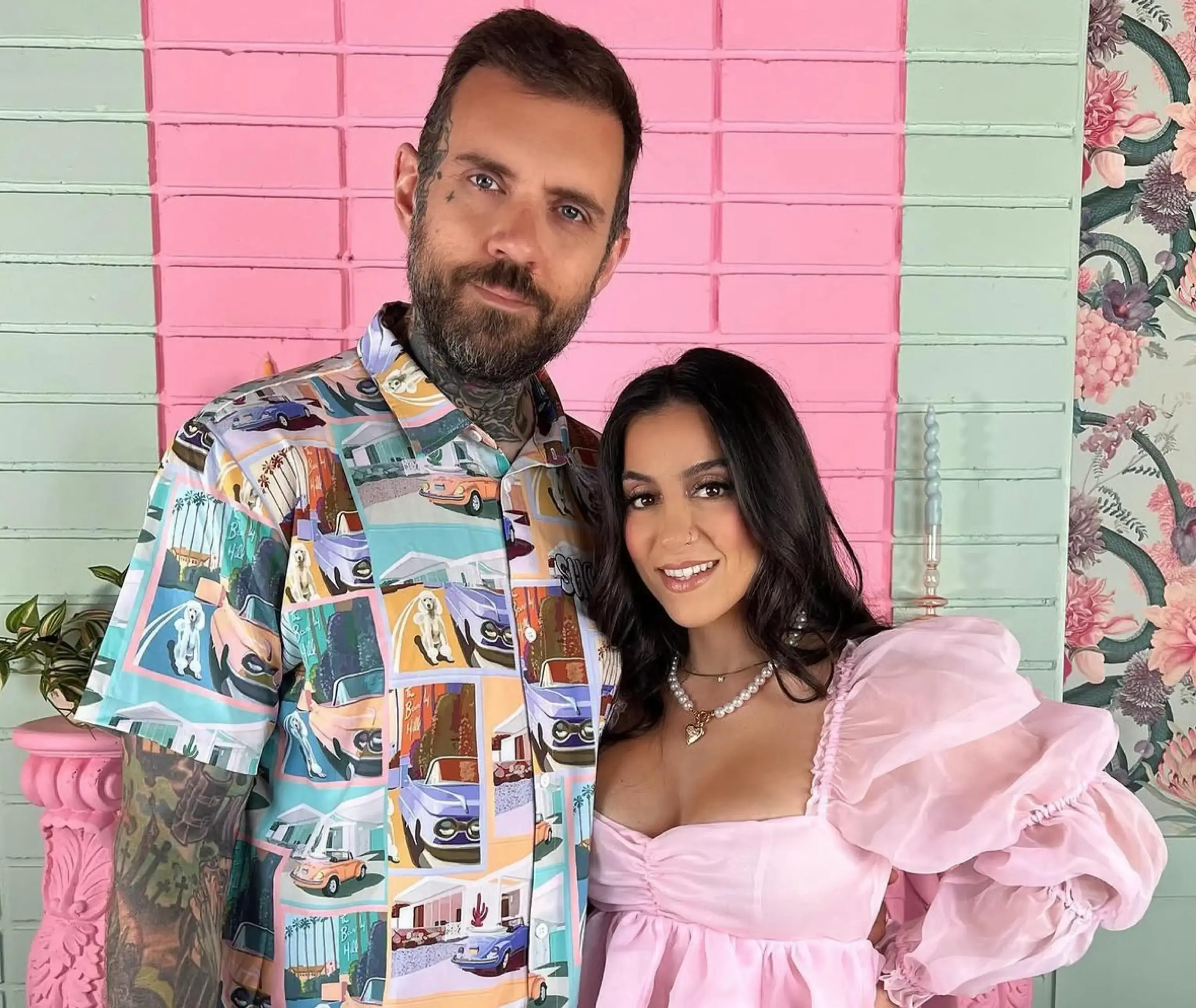 Adam22, wearing a colorful car-print button-up shirt, stands beside Lena the Plug in a puffy pink dress against a pink and mint green brick background