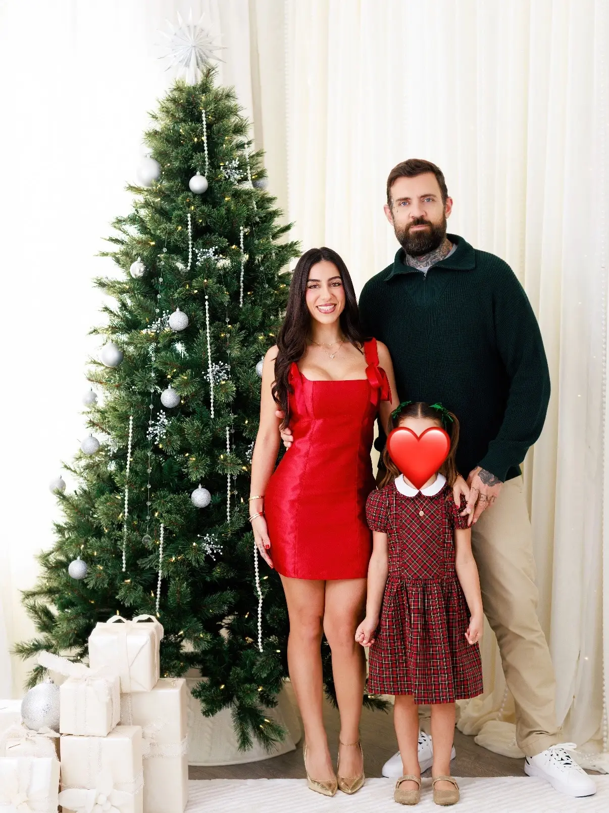 Adam22 and Lena stand with their young child, whose face is covered by a red heart emoji, next to a decorated Christmas tree