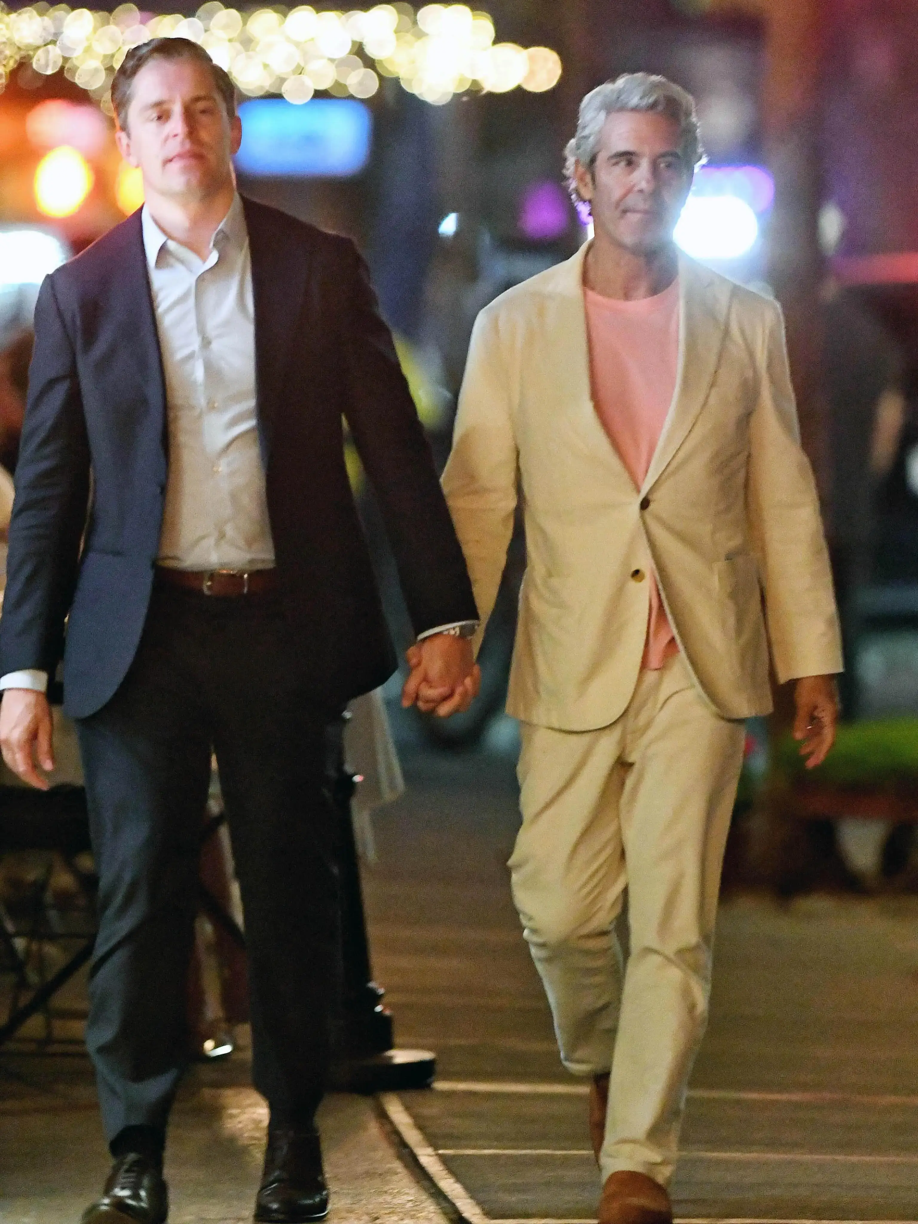 Andy Cohen in a cream suit holds hands with Kevin Sobieski in a dark suit while walking together at night.