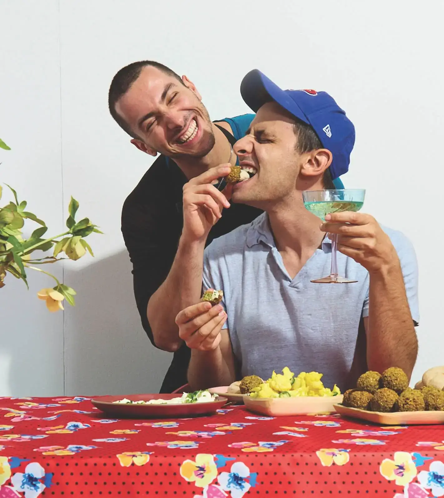 Benj Pasek laughs from behind while playfully feeding a piece of food to Jake Cohen, who holds a cocktail glass at a dining table