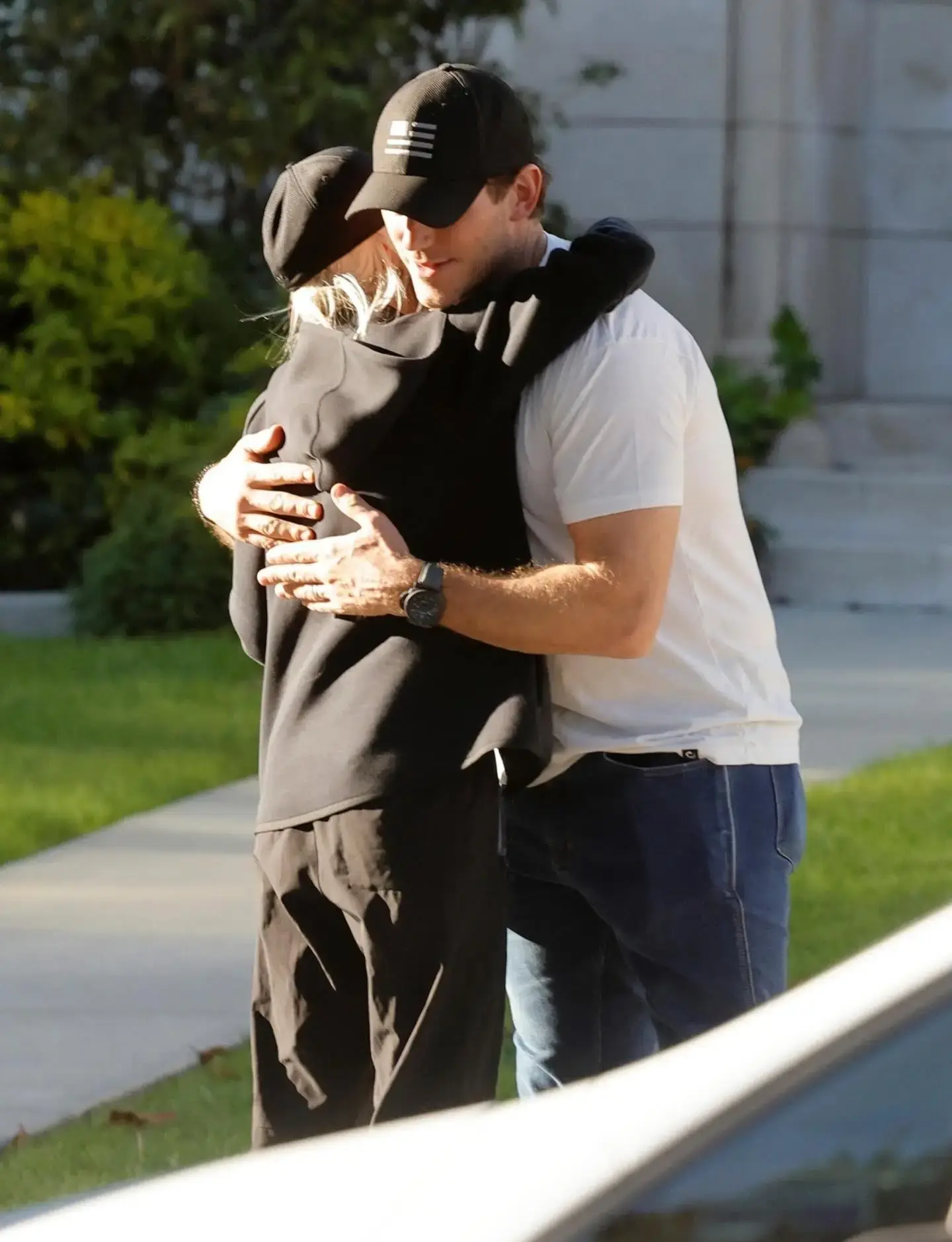 Chris Pratt in a white t-shirt and baseball cap embraces Anna Faris, who is wearing an all-black casual outfit and hat, outside on a sidewalk.