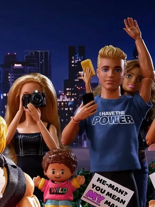 A group of Mattel toy figures, including Ken in an "I Have The Power" shirt and a Barbie doll holding a camera, gather against a night city skyline backdrop