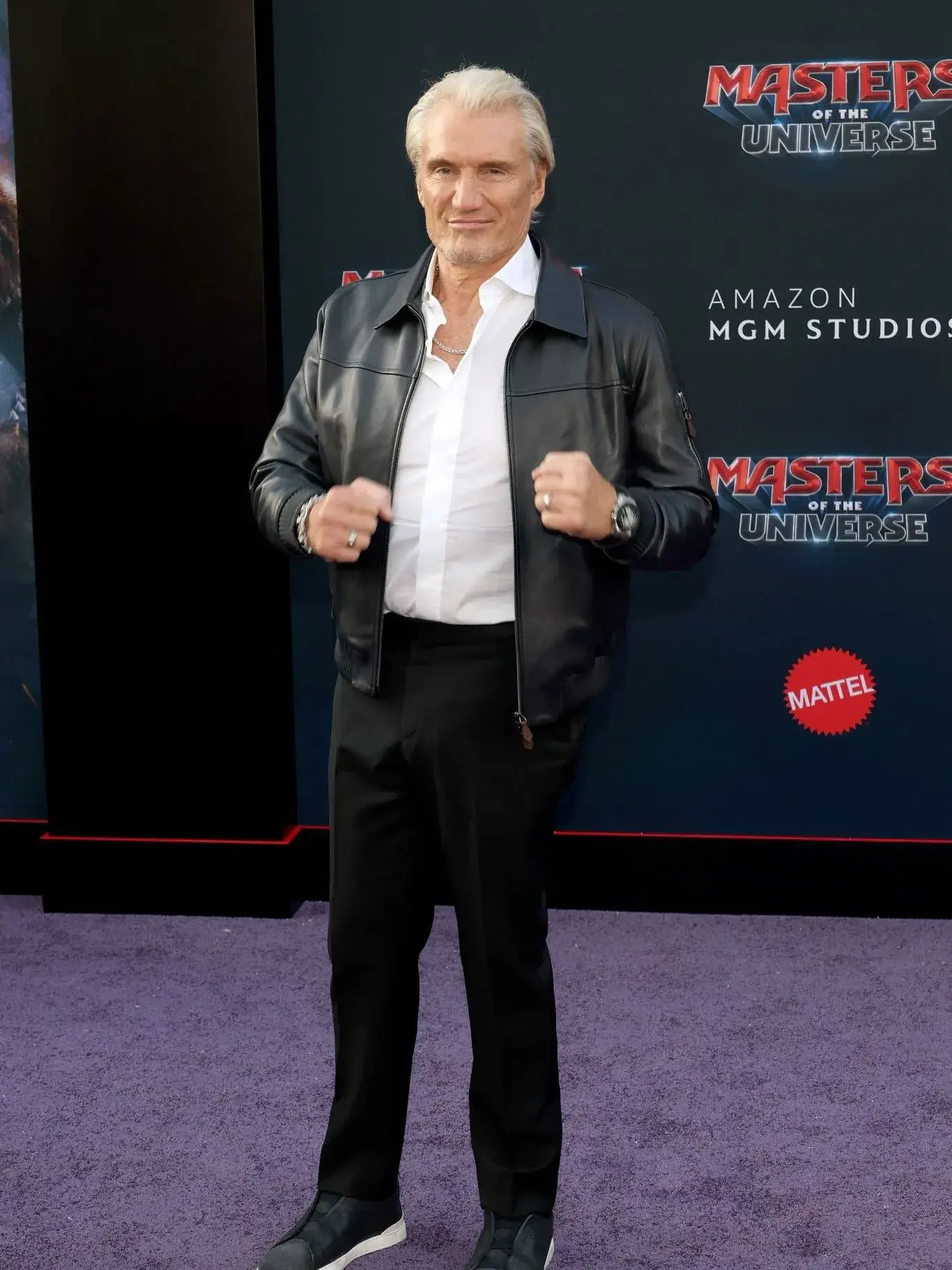 Actor Dolph Lundgren poses on a purple carpet wearing a black leather jacket, white button-down shirt, and black trousers.