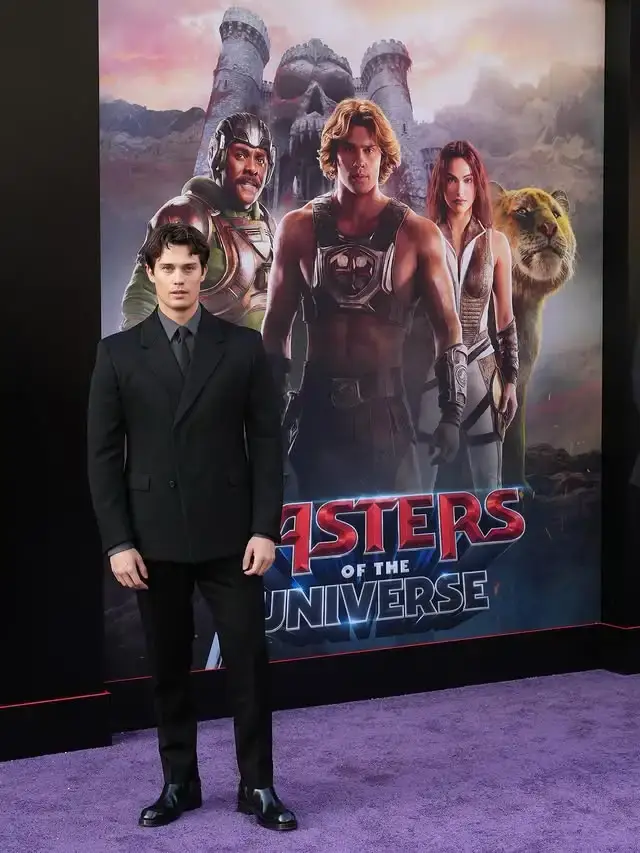 Nicholas Galitzine stands in a sharp black suit on a purple carpet in front of a Masters of the Universe movie poster.
