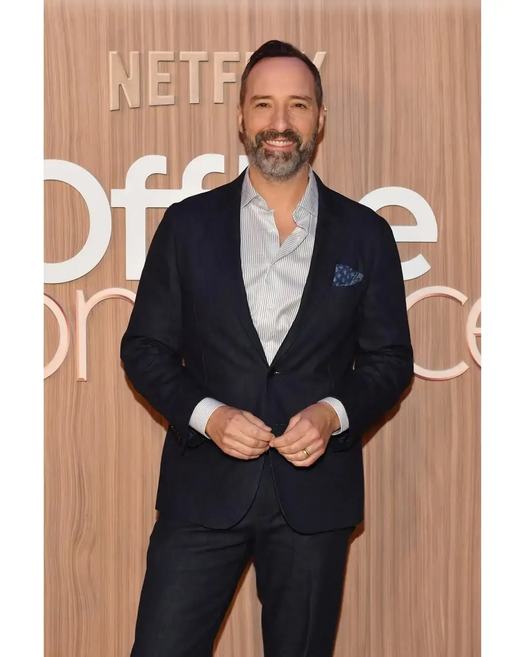 Actor Tony Hale smiles while posing on a red carpet in a dark blazer, pinstriped button-down shirt, and patterned pocket square against a wood-paneled Netflix backdrop.