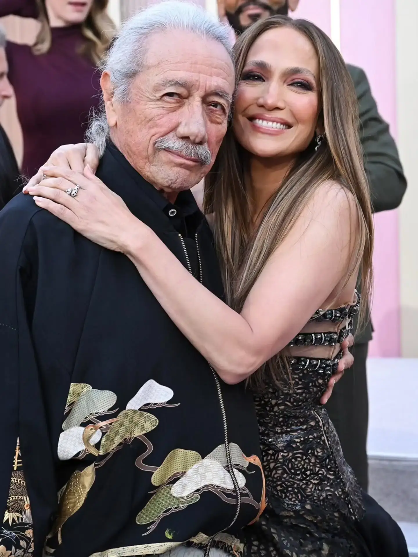 Jennifer Lopez embraces Edward James Olmos on a red carpet while he wears a black jacket with a golden crane pattern