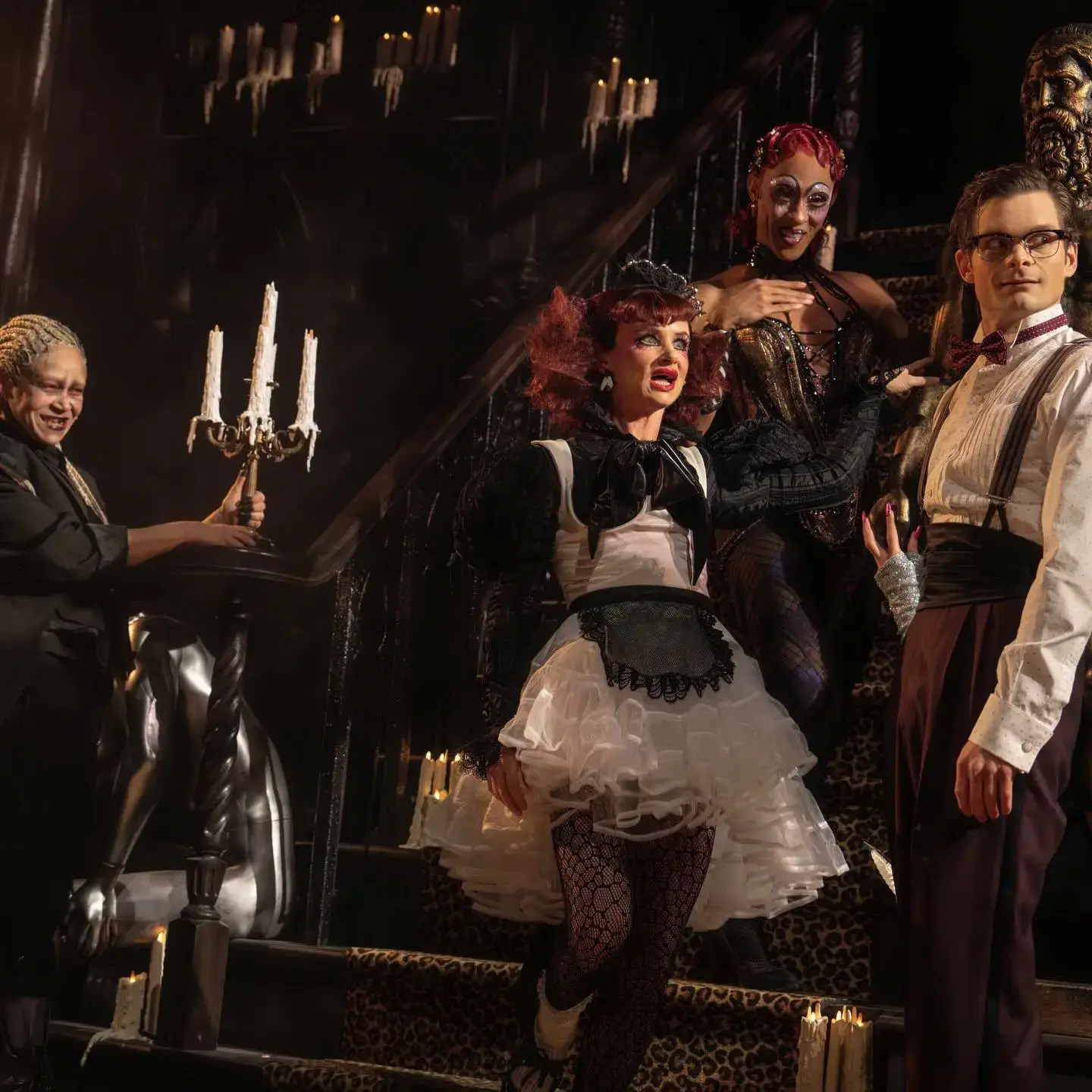 Cast members from The Rocky Horror Show perform on a dimly lit, Gothic staircase surrounded by melting candles