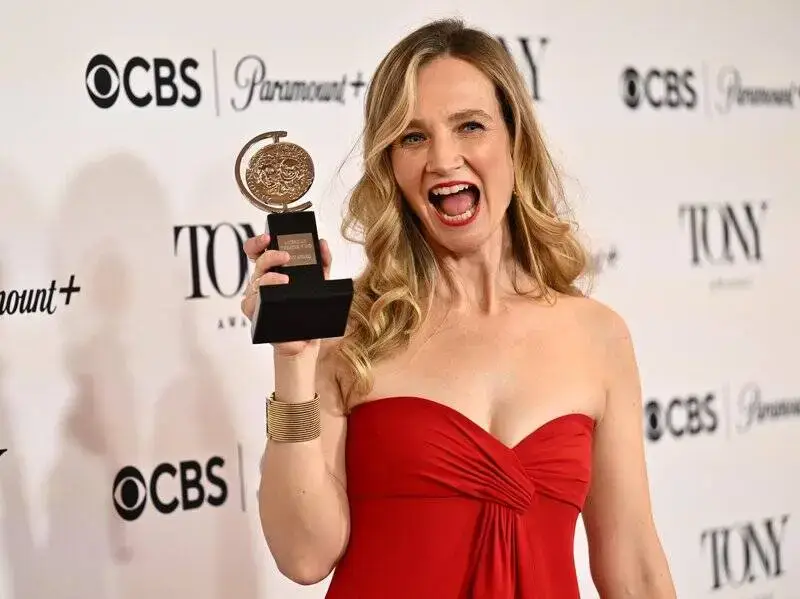 Bess Wohl in a red strapless dress smiles excitedly while holding up a Tony Award in front of a branded press backdrop.