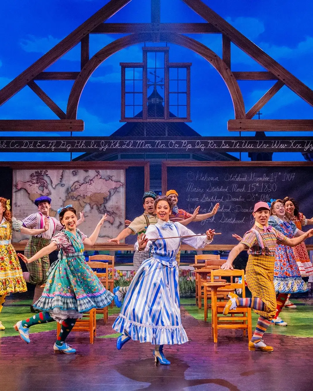 Cast members perform a lively, coordinated dance routine in colorful vintage attire on a schoolhouse-themed stage set