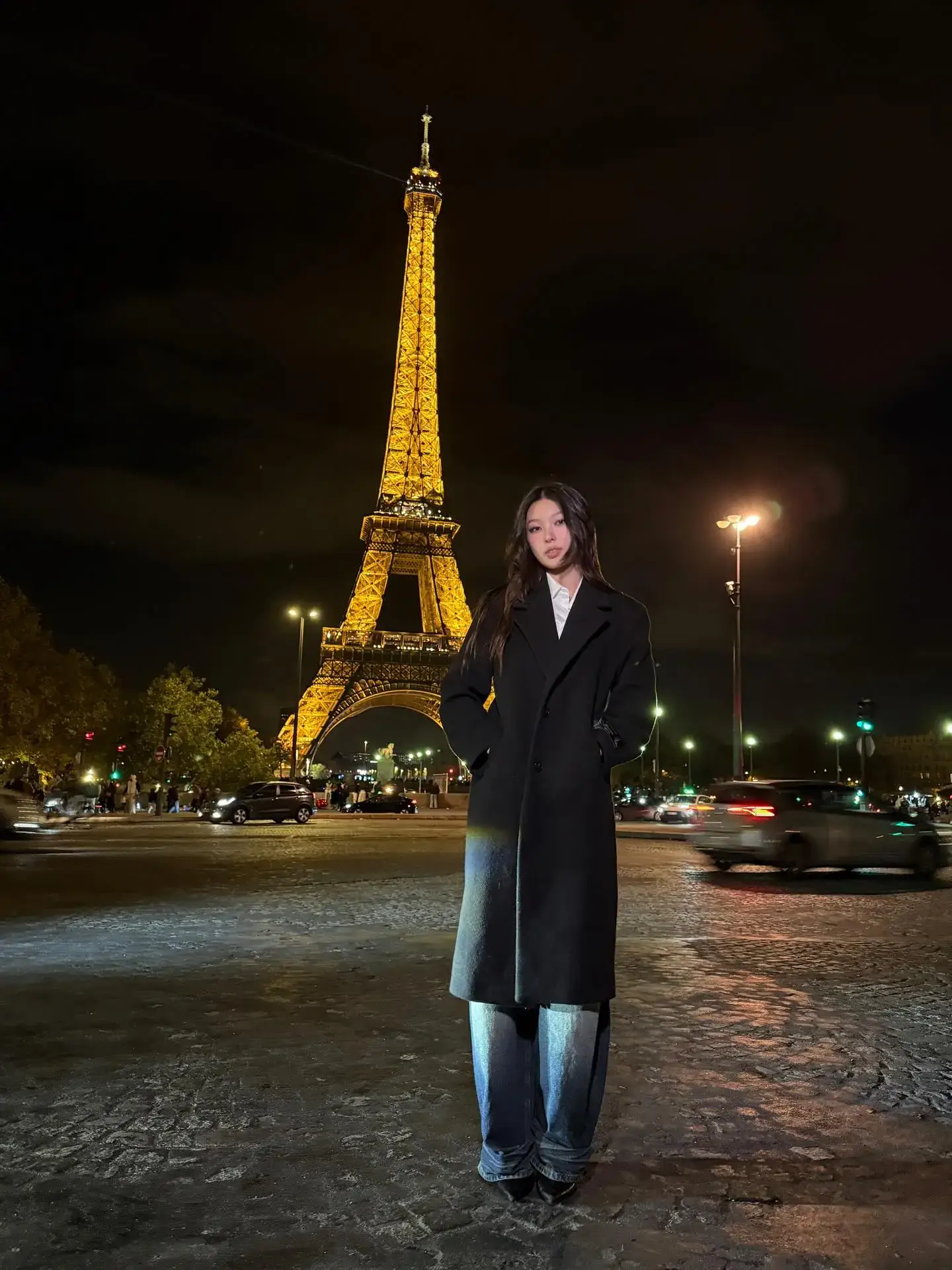 Yoonchae stands in a long black winter coat and wide-leg jeans with the illuminated Eiffel Tower glowing behind her at night.