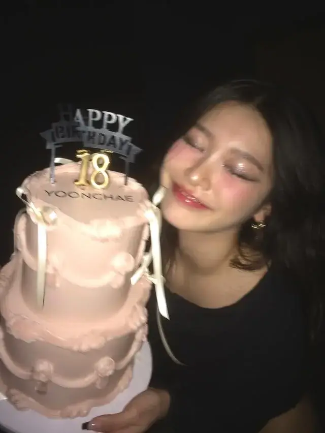 Yoonchae smiles happily with her eyes closed as she poses next to a three-tiered pink 18th birthday cake.