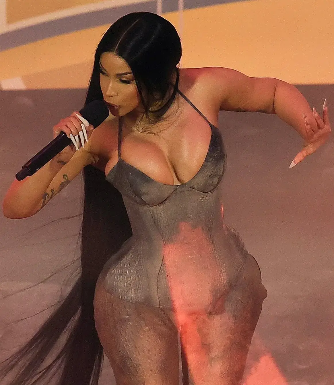 Cardi B sings into a microphone with long flowing hair, wearing a form-fitting grey textured halter jumpsuit under stage lighting.