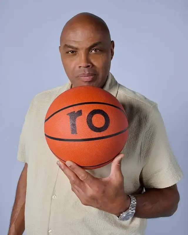 Charles Barkley holds an orange basketball printed with the letters "ro" while wearing a beige short-sleeved button-down shirt.