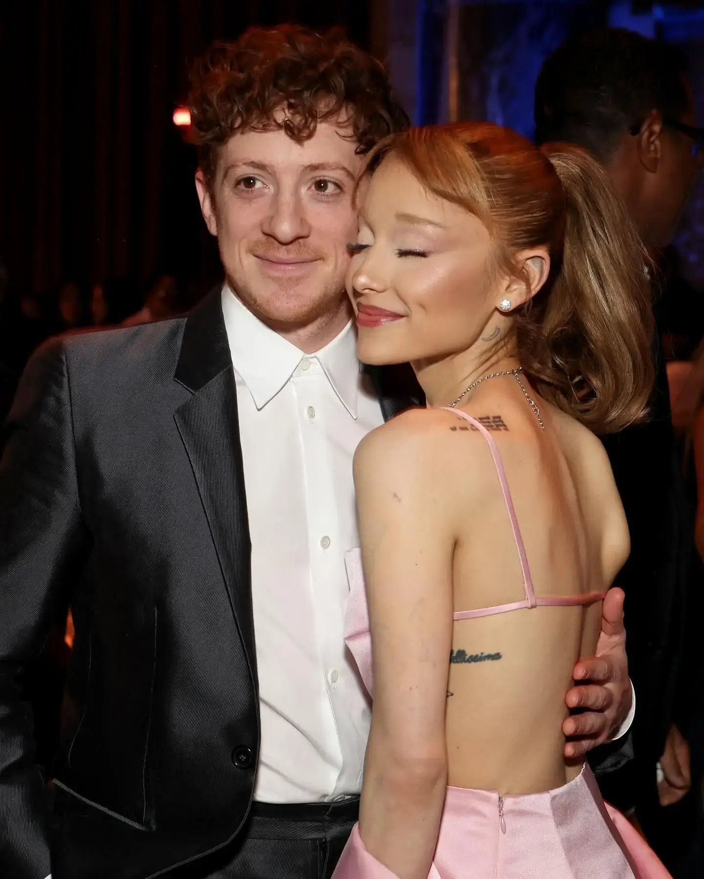 Ethan Slater in a dark suit jacket and white button-down shirt embraces Ariana Grande, who smiles with her eyes closed in a strappy pink gown.