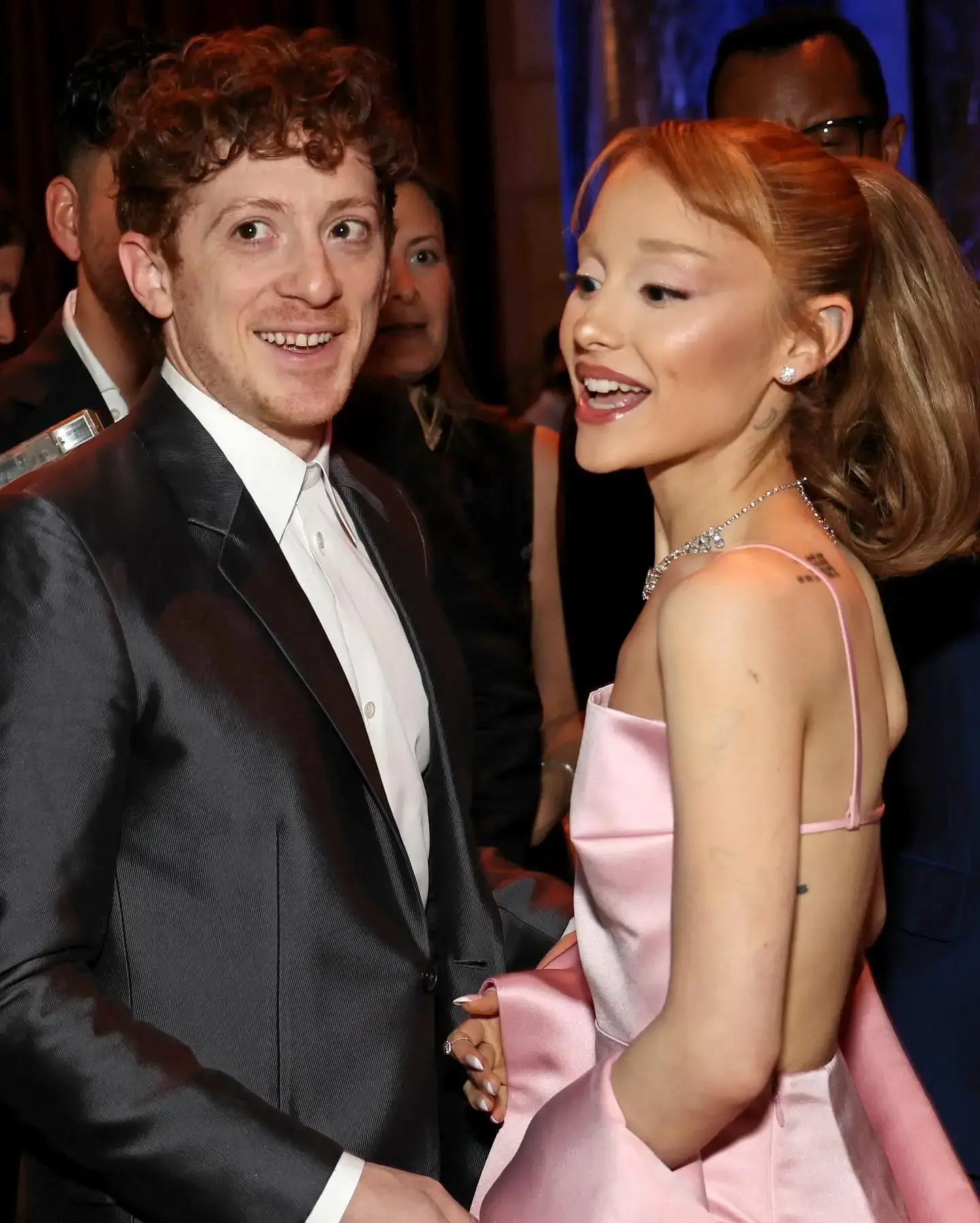 Ethan Slater in a dark suit jacket stands next to Ariana Grande, who is talking and smiling in a strappy pink gown