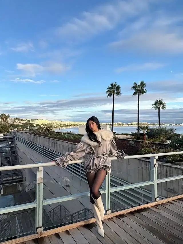 Sophia Laforteza stands on a wooden balcony railing wearing a grey romper with a fur collar and tall white boots against a coastal city background.