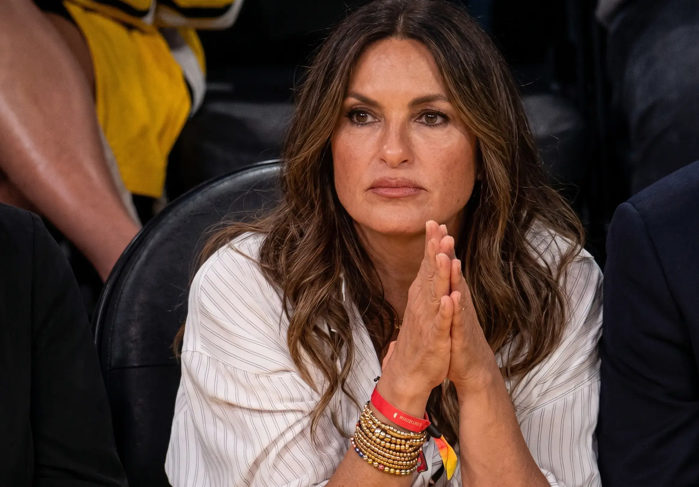Mariska Hargitay sits courtside in a white pinstripe button-down shirt with her hands clasped together.